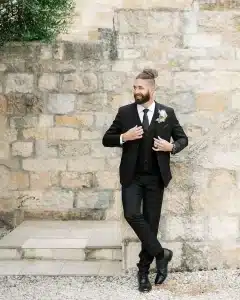 Groom posing against a wall in a classic black wedding suit, black pants, black vest, black tie, pocket square, and white shirt 