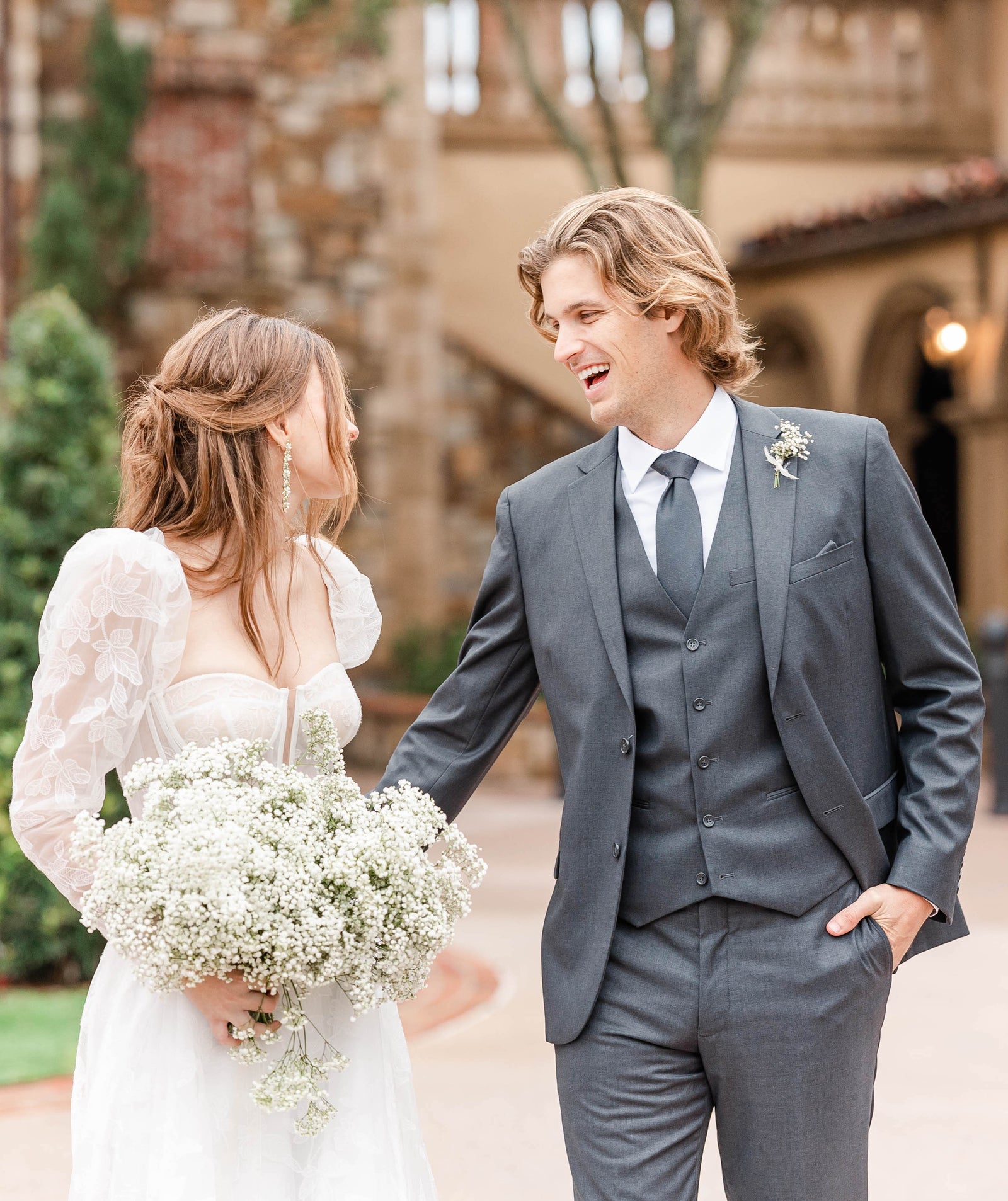 A bride in a white wedding dress holding flowers looking back at her groom wearing a light grey suit