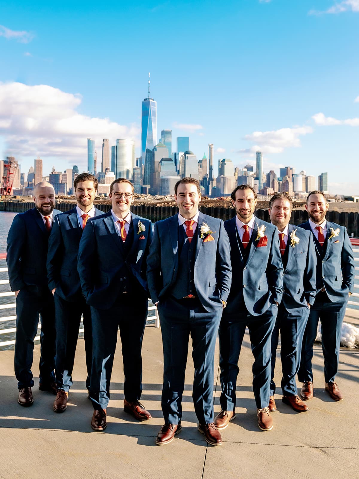 A group of men in navy wedding suits stands in a v-formation with skyscrapers in the background.