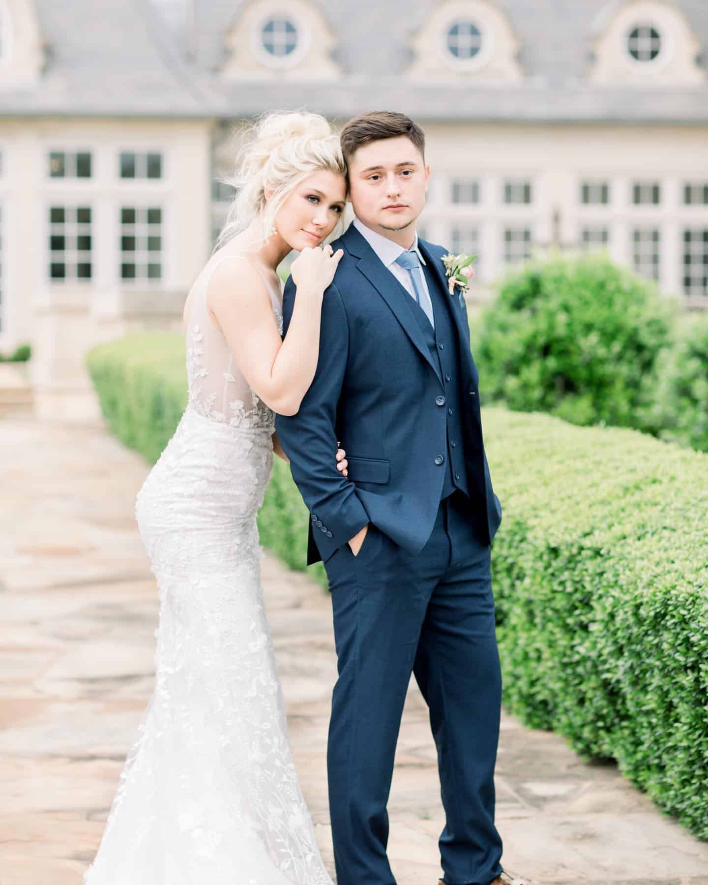 A groom in a navy blue suit stands with his bride resting her hands and head on his shoulder.