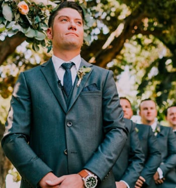 Men wearing charcoal-gray wedding suits pose together under a tree.