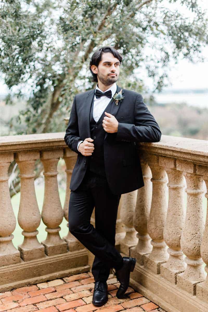 A groom in a black suit stands on a bricked balcony.