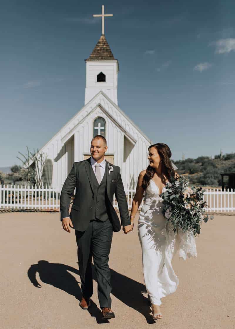 A man in a grey wedding suit poses with his bride in front of a church.