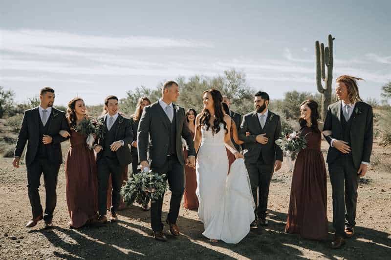 A bride and groom pose with their bridal party dressed in black suits, burgundy dresses, and accessories.