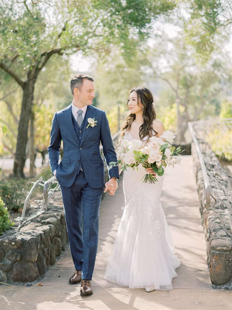 The bride and groom hold hands as they walk down a tree-covered path.