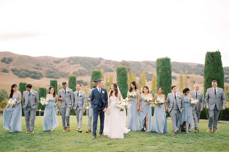 A shot of a bride and groom with their bridal party dressed in gray and light blue, walking outdoors.