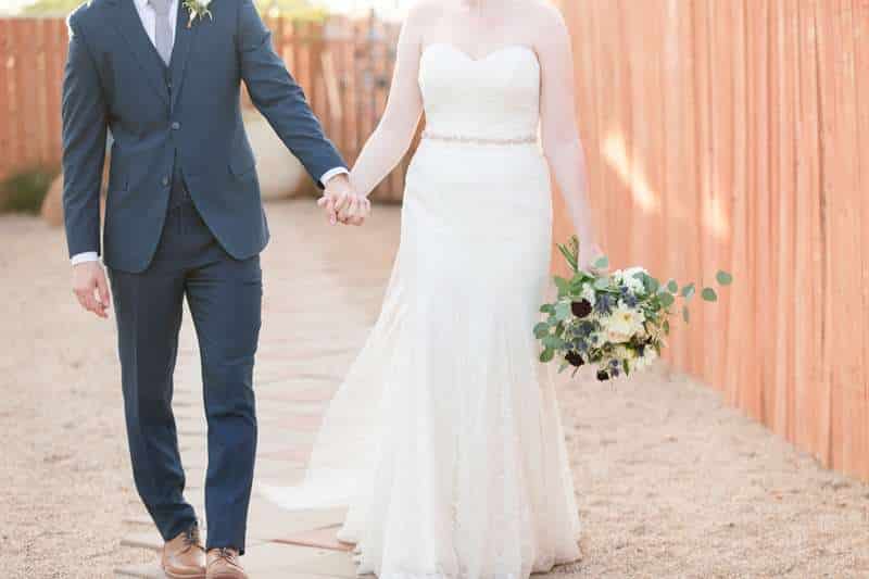 A newlywed couple holds hands as they walk down a gravel walkway.