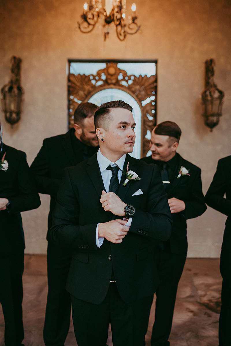 Man in a black modern groom suit adjusting his cuff in front of his groomsmen.