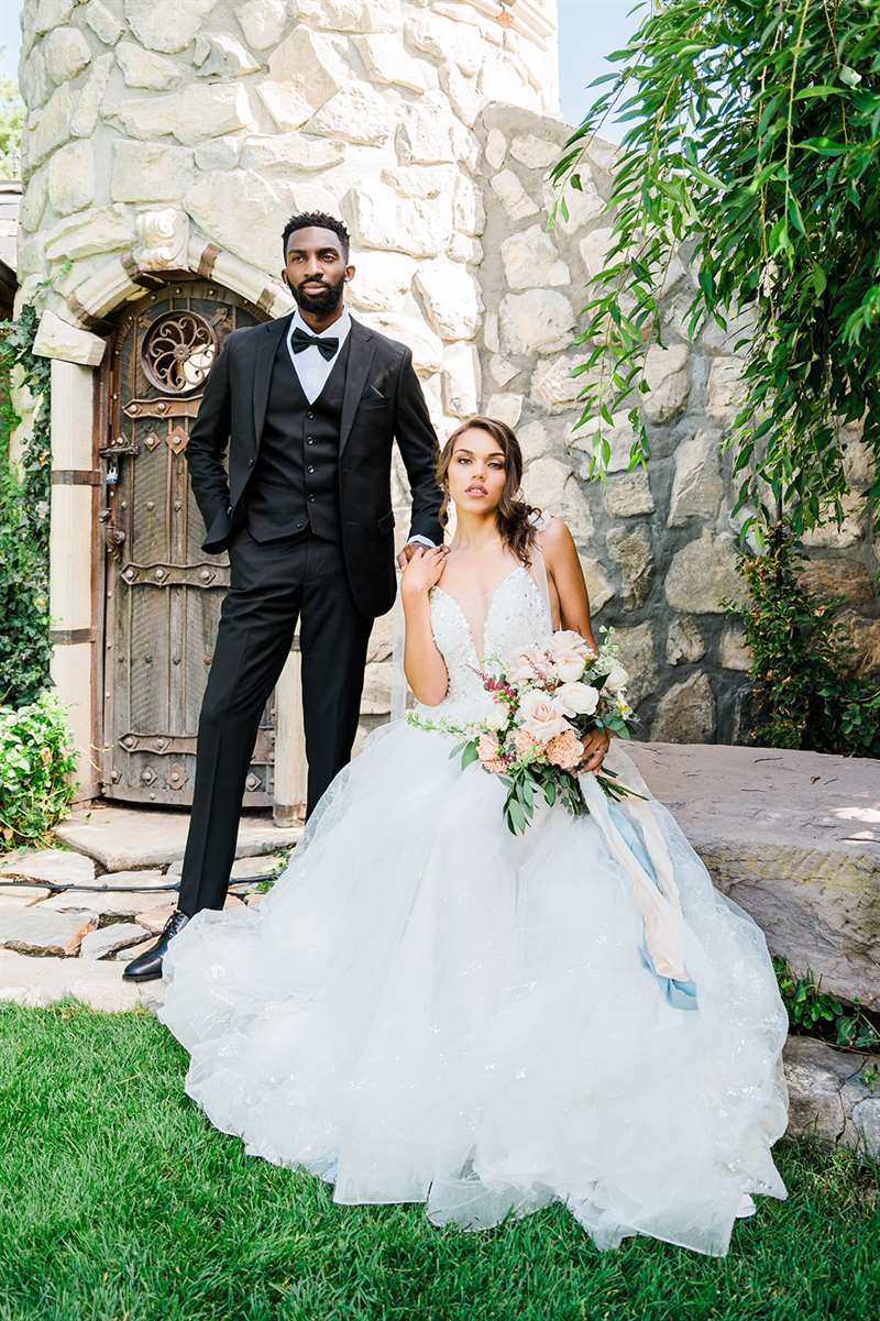 A man in a black wedding suit poses next to his bride in a garden.