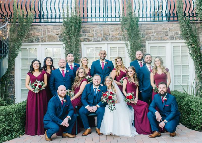 A bride and groom pose with their bridal party, dressed in blue suits and burgundy dresses and accessories.