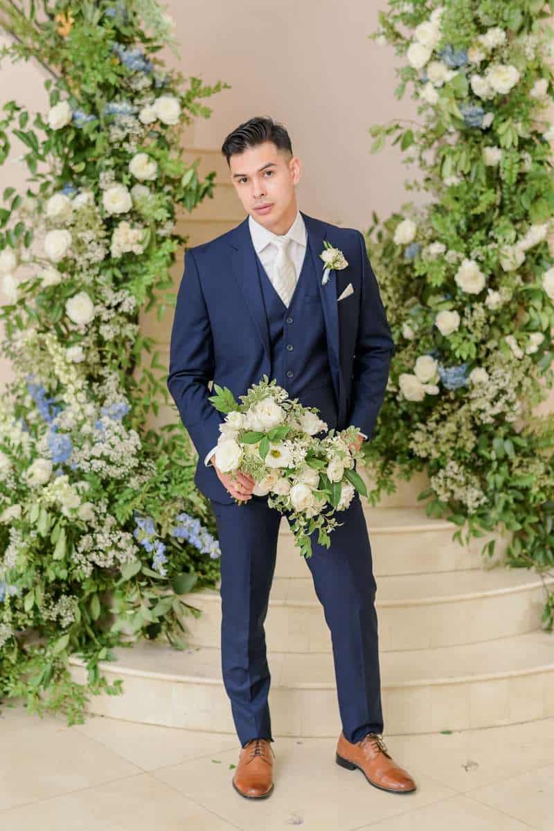 Groom in a dark blue suit posing with a bouquet and flowers behind him.