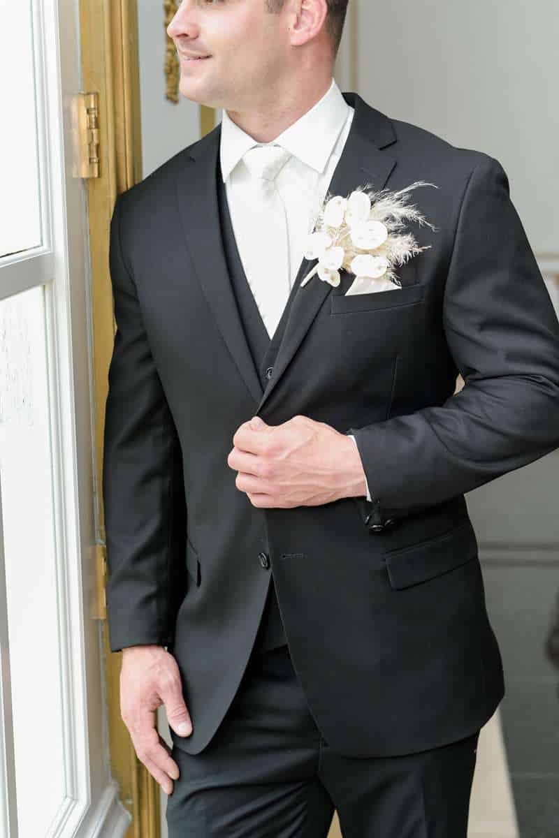 A man looks out the window wearing a black suit and boutonniere.