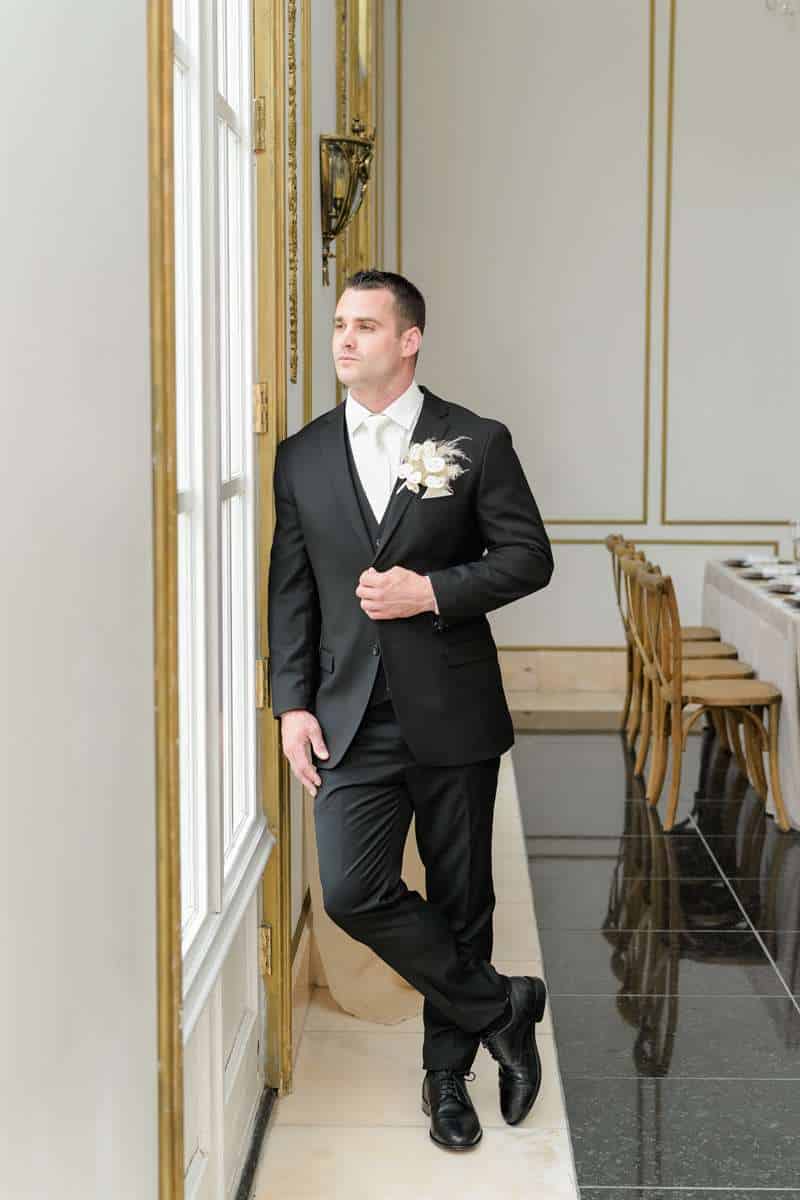 A man in a black, Modern Groom suit stands next to a window in a reception hall.