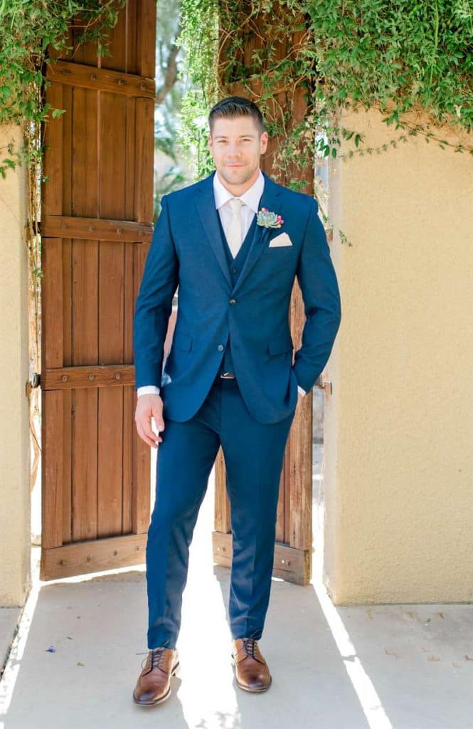 A man standing by a door poses in a blue three-piece blue wedding suit and boutonniere with cognac shoes.