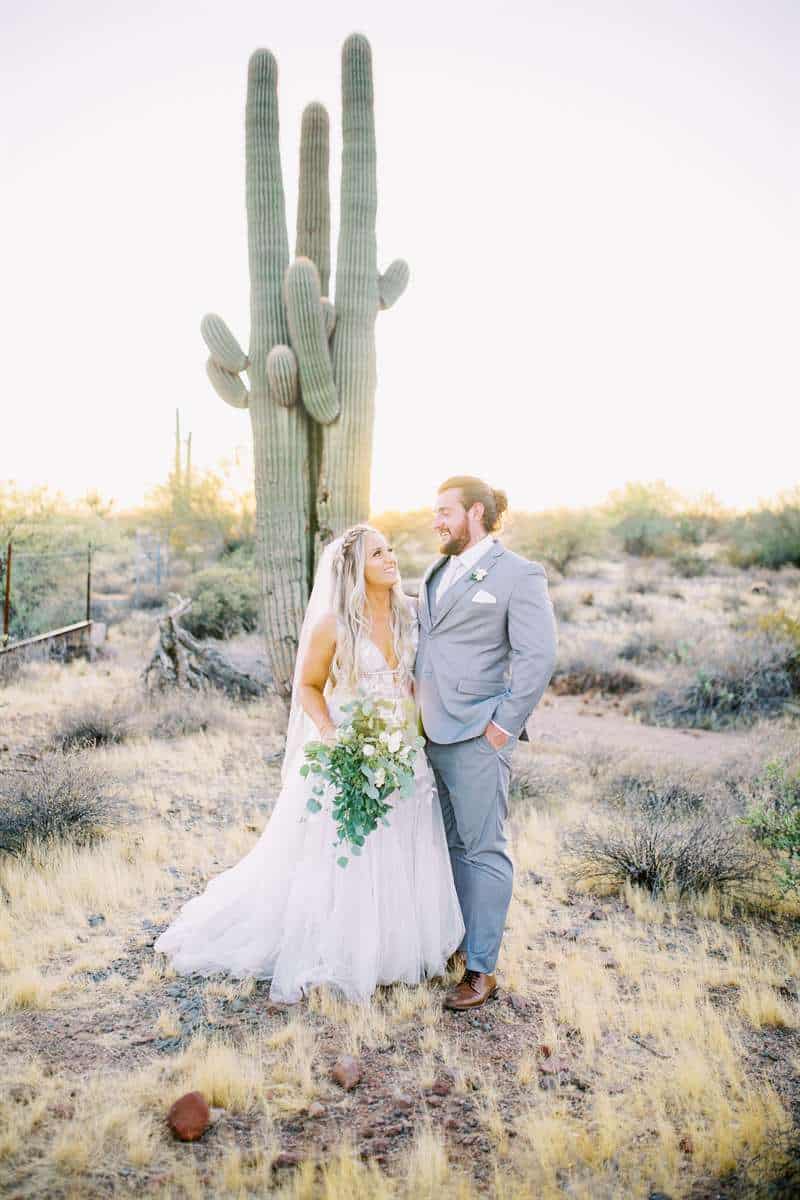 A bride and groom pose by a cactus. The groom wears a light gray suit.