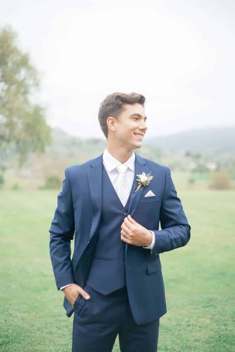 A man in a blue wedding suit smiles while standing in a spacious pasture.