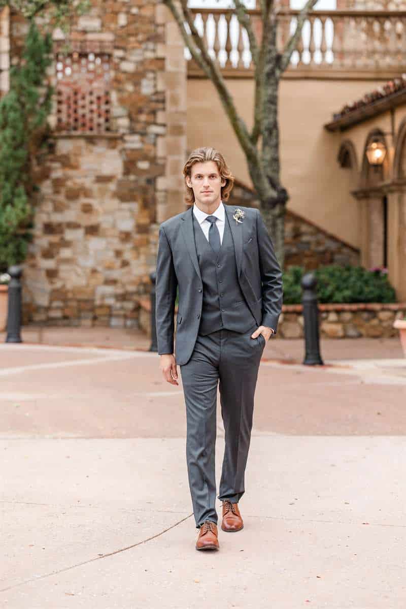A male model walks towards the camera wearing a grey groom suit in front of a wedding venue.