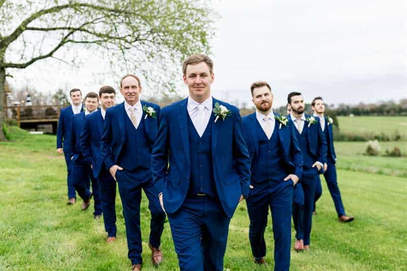 The groom leads the groomsmen in a v-shape pattern across a green field in navy blue suits.