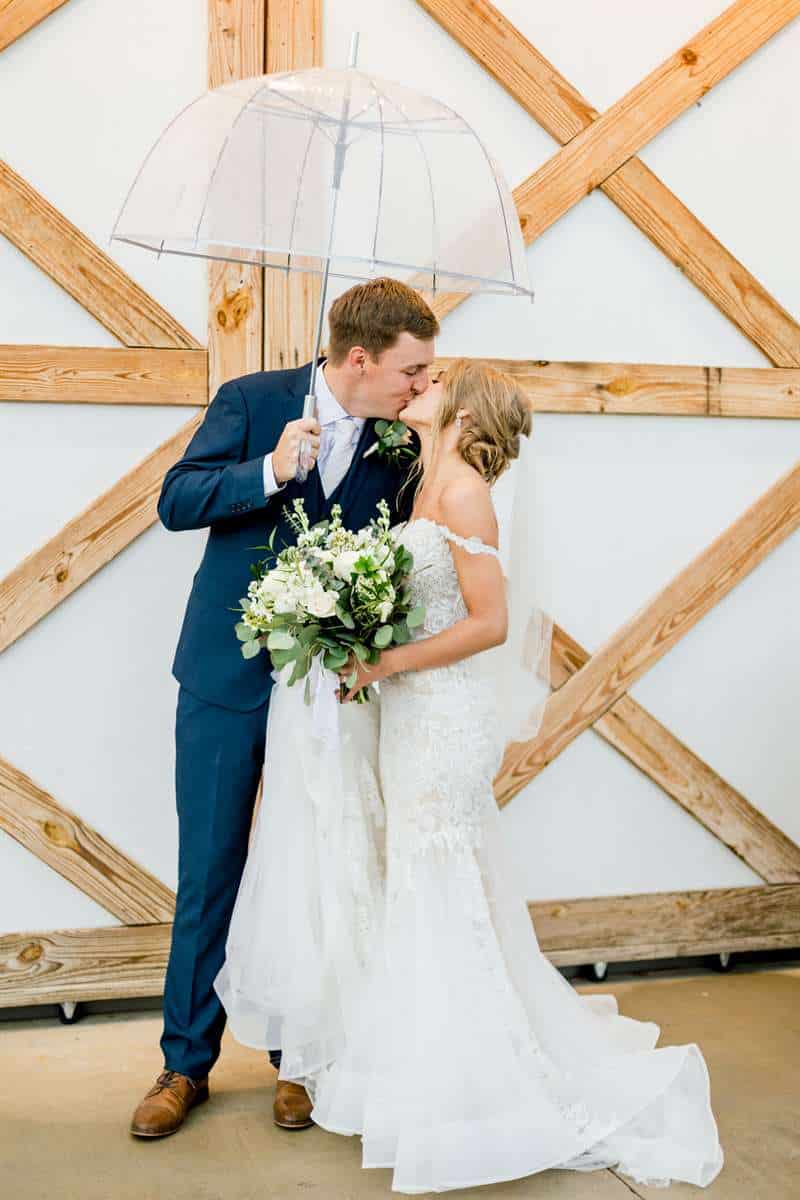 A bride and groom kiss under an umbrella. The groom wears a navy blue suit.