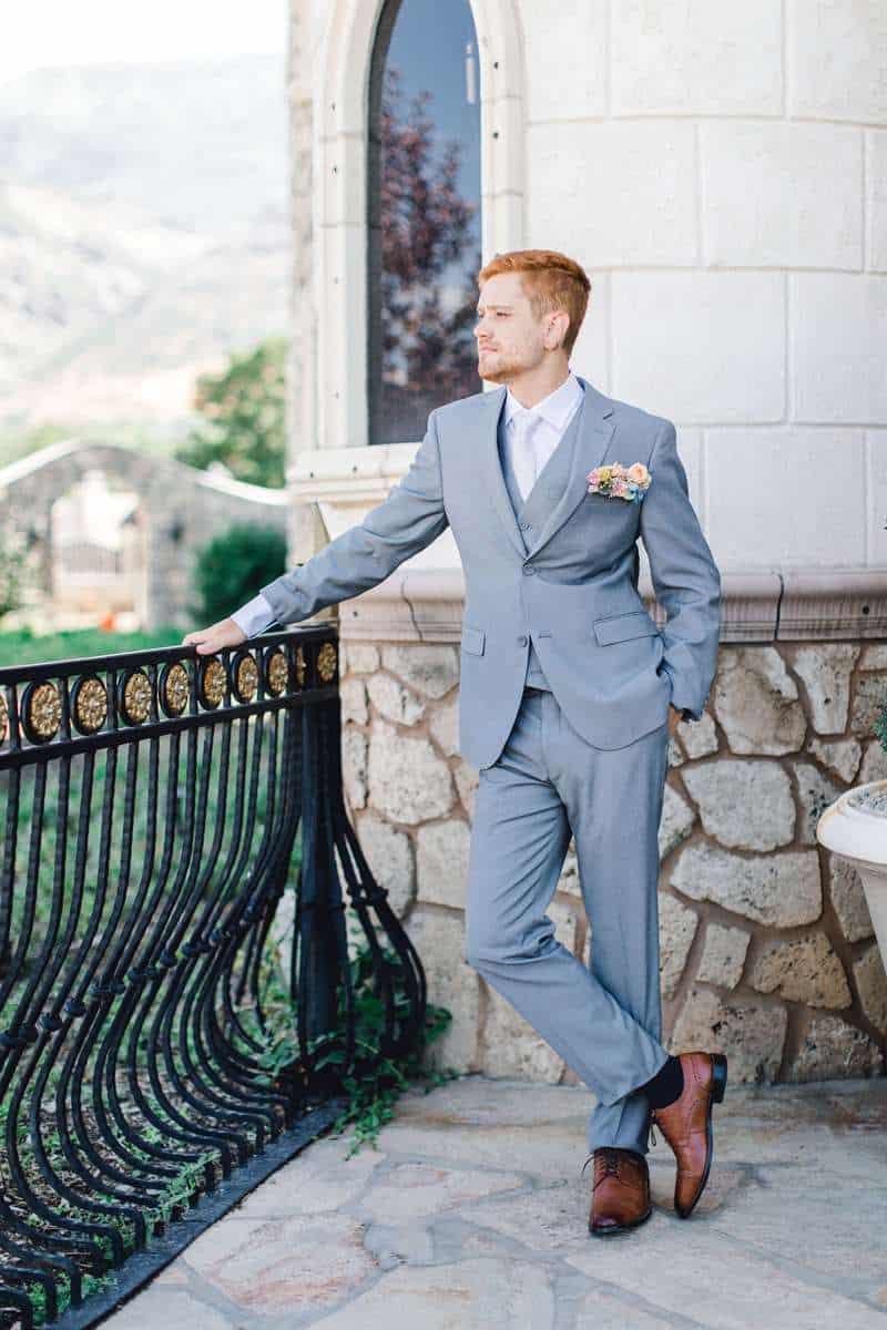 A male model poses on a balcony with a light grey suit and boutonniere.