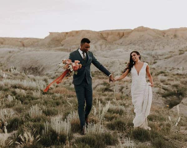 A young newlywed couple holds hands in the middle of a desert field landscape.