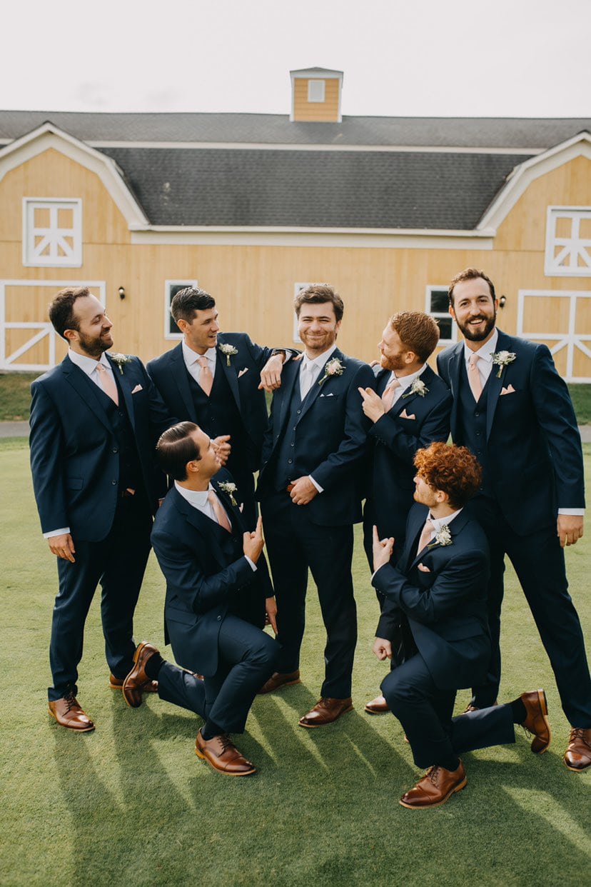 Groomsmen looking at a smiling groom asking who pays for groomsmen suits in front of a yellow barn