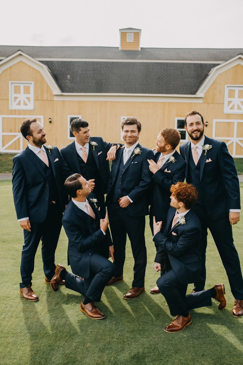 Groom with groomsmen surrounding him wearing black wedding suits in front of a yellow barn