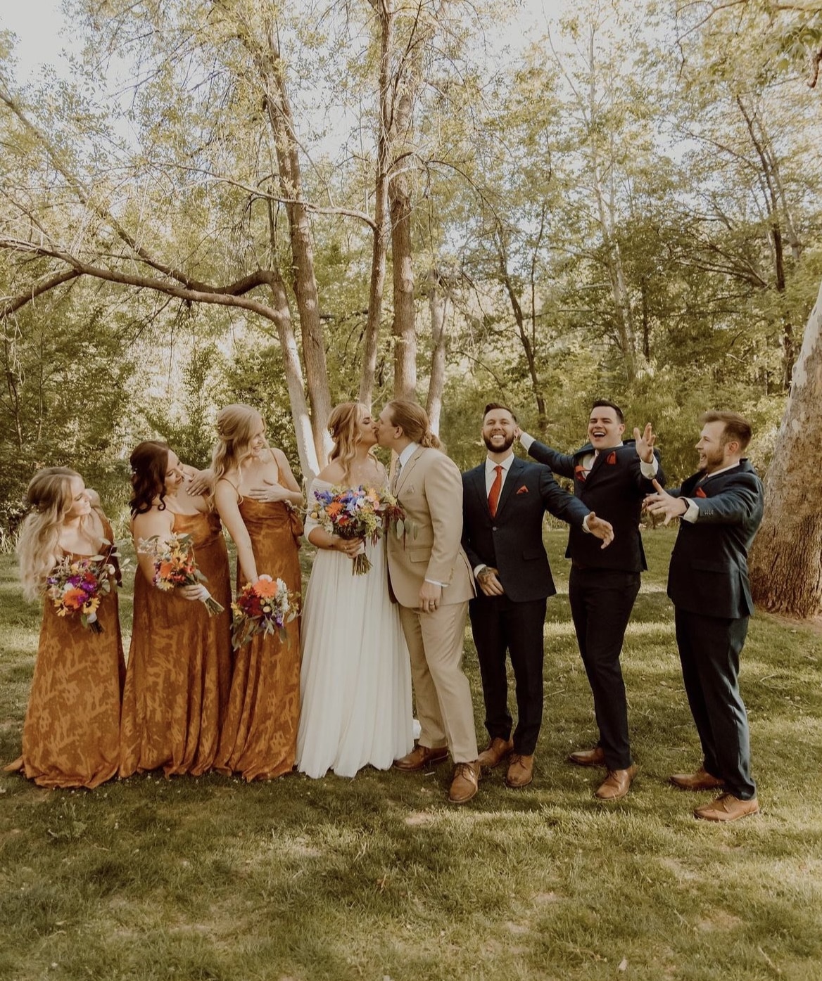Groom in a tan summer wedding suit kisses his bride while the bridal party watches and cheers