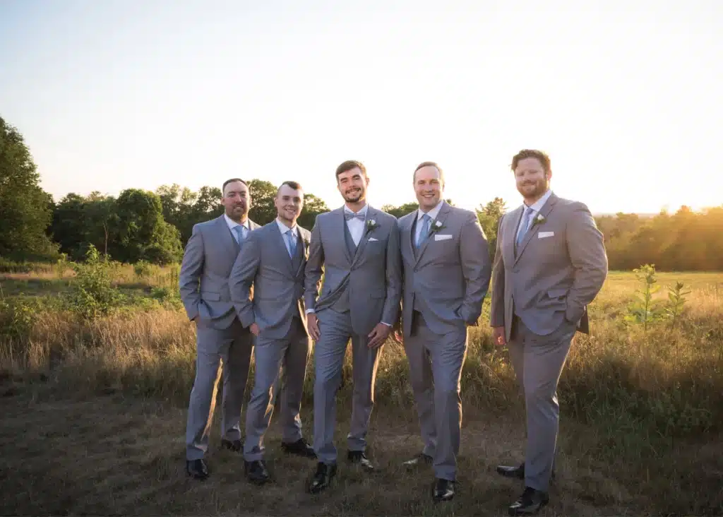 Groom and groomsmen in light grey suits with matching black shoes standing in a field at sunset