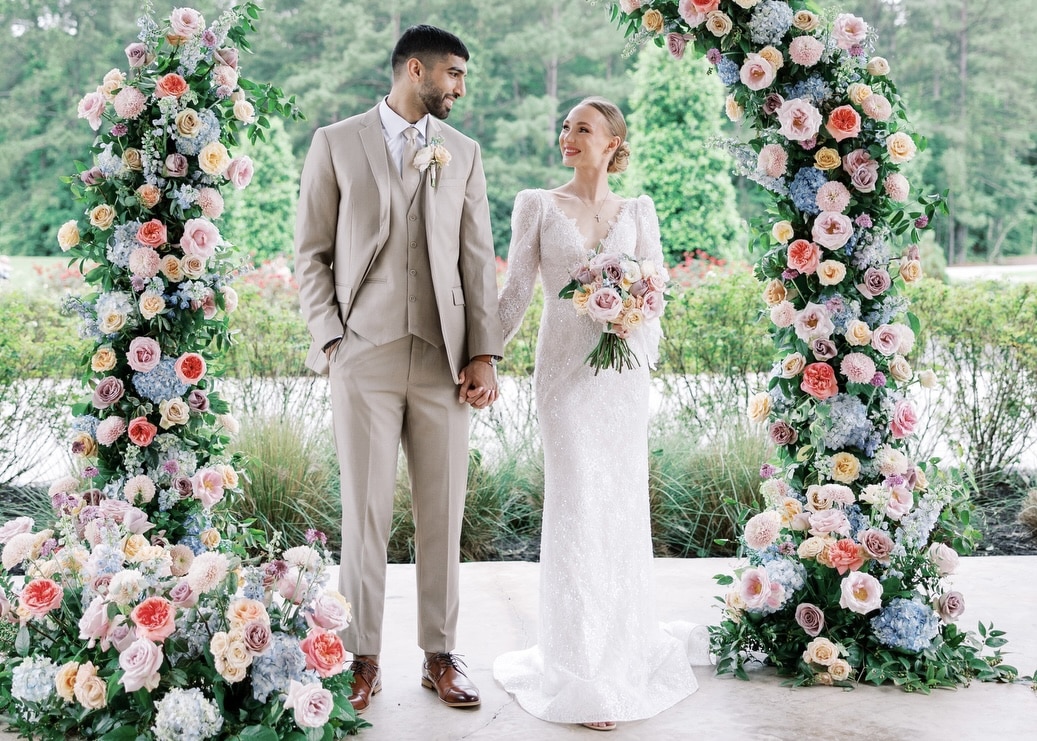 A groom wearing a summer wedding suit stands with his bride under a floral arch outside