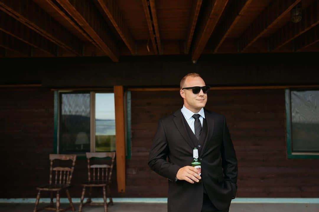 Groom holding a beer wearing a black wedding suit and black shades on the porch of a cabin