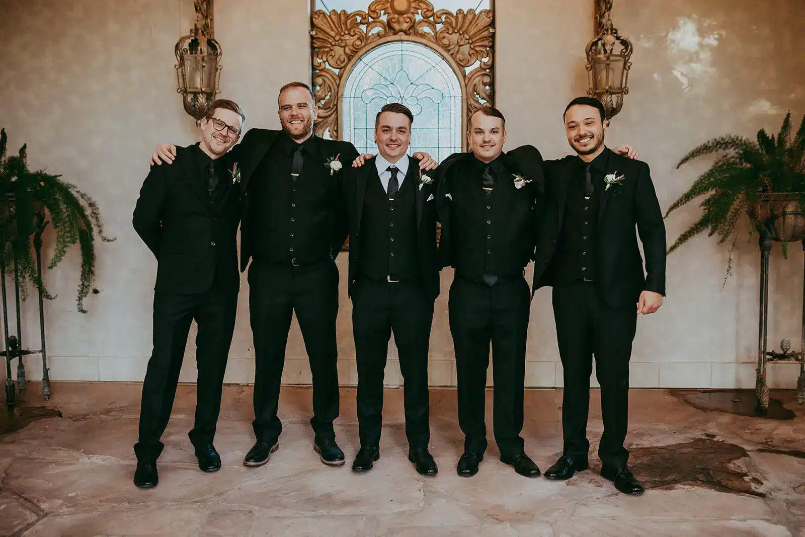 Groom and groomsmen lined up against a wall with arms around each other’s shoulders wearing black-tie wedding suits