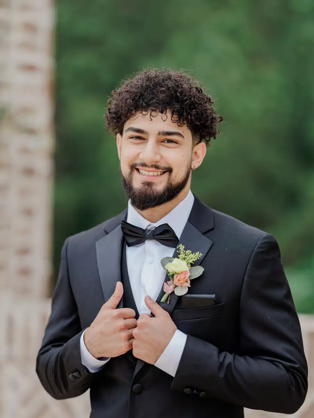 Man with curly hair smiling outside while holding on to the lapels of his black wedding tuxedo for a black-tie wedding