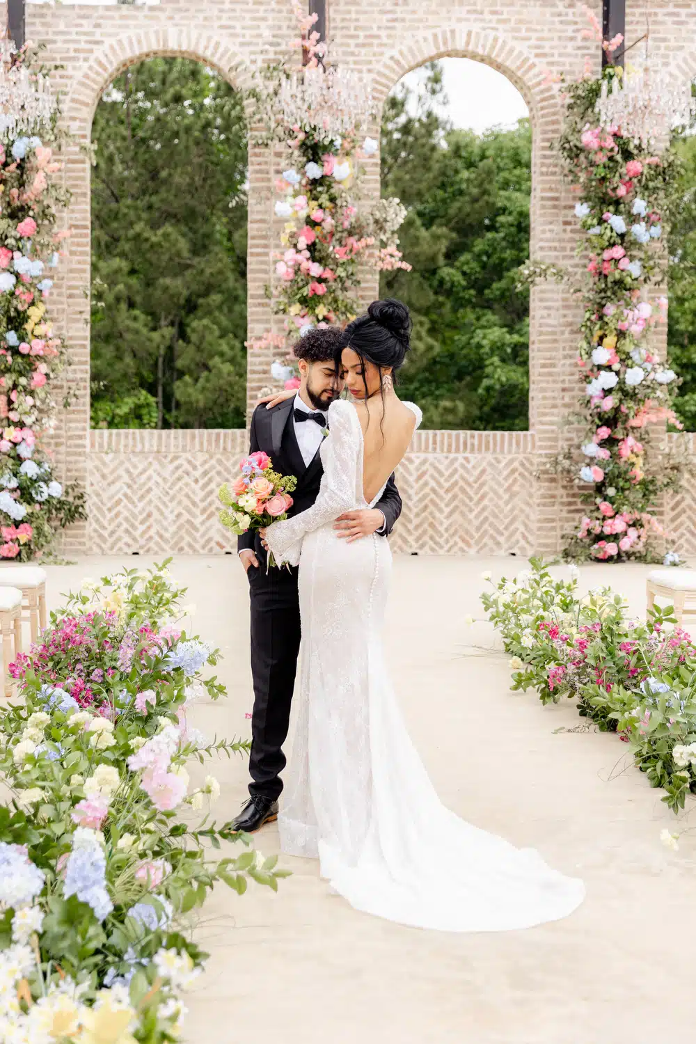 Groom wearing a black-tie wedding tuxedo holding a bride wearing a wedding gown outdoors in a colorful garden of flowers
