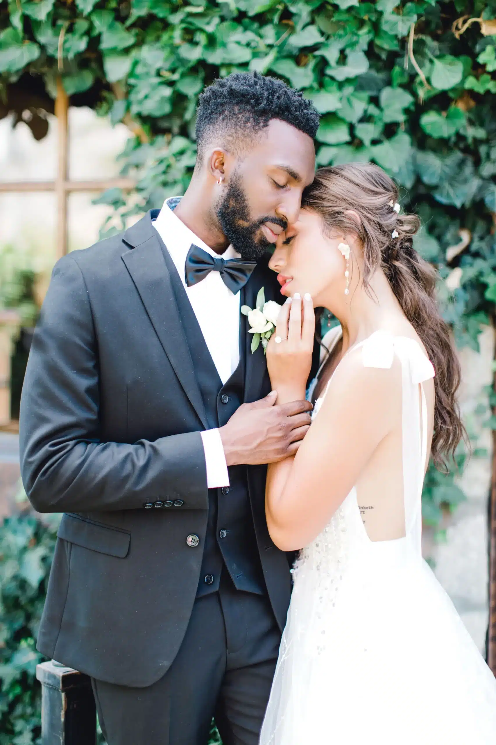 Groom wearing a black-tie wedding suit and bride in her wedding dress holding each other with their eyes closed