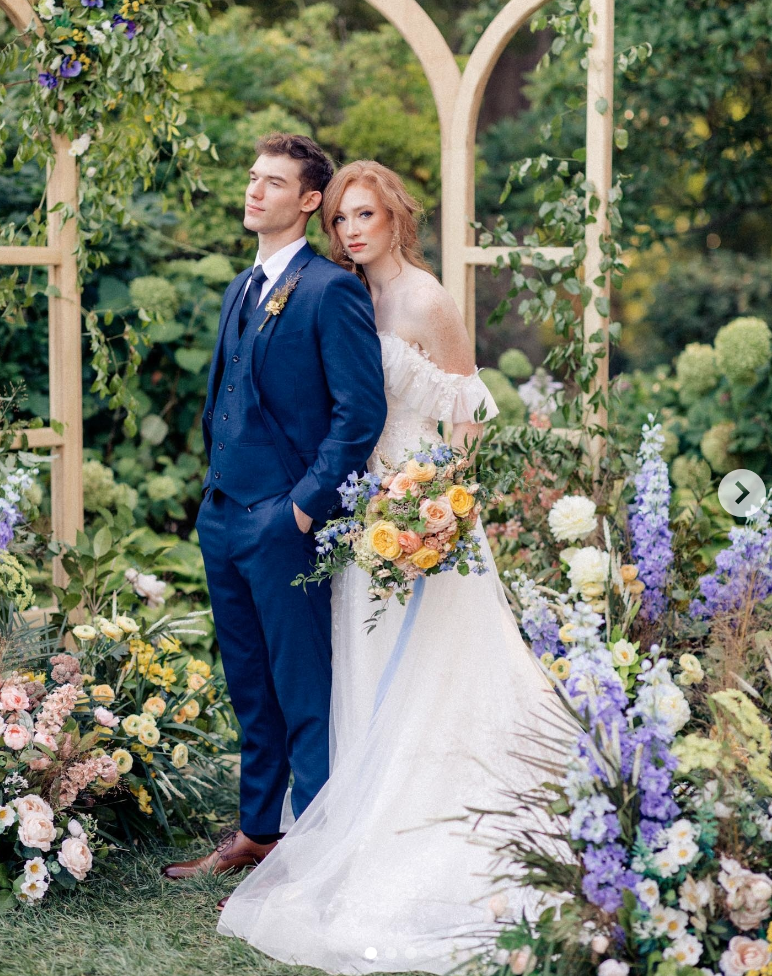 Bride holding the arm of a groom wearing a midnight blue wedding suit in a courtyard of flowers