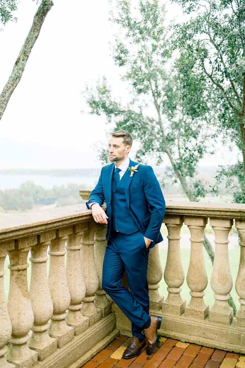 Alt text: Groom in semi formal wedding attire poses against a stone balustrade